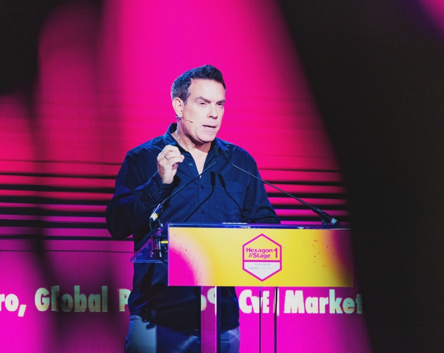 Man with short dark hair wearing a dark blue shirt speaking at a podium with a bright pink and yellow Hexagon Stage banner in the background.