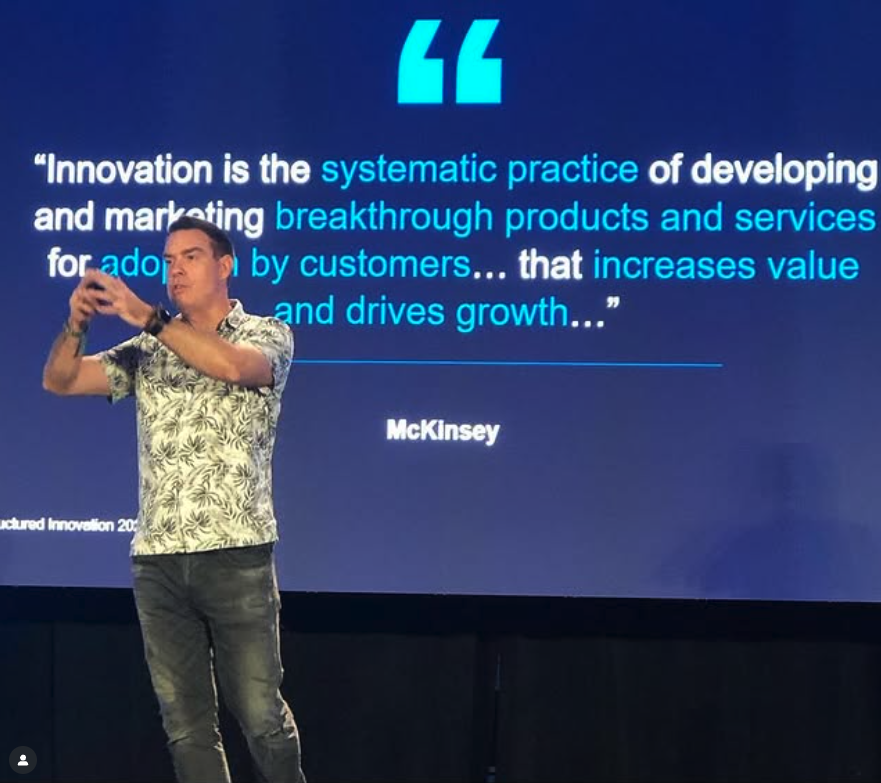 Man in a patterned short-sleeve shirt and jeans speaking on stage with a large screen behind displaying a McKinsey quote about innovation as systematic practice increasing value and driving growth.