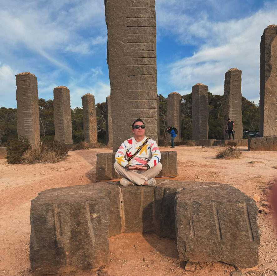 Man sitting cross-legged on a stone bench in front of tall carved stone pillars outdoors under a blue sky with scattered clouds.