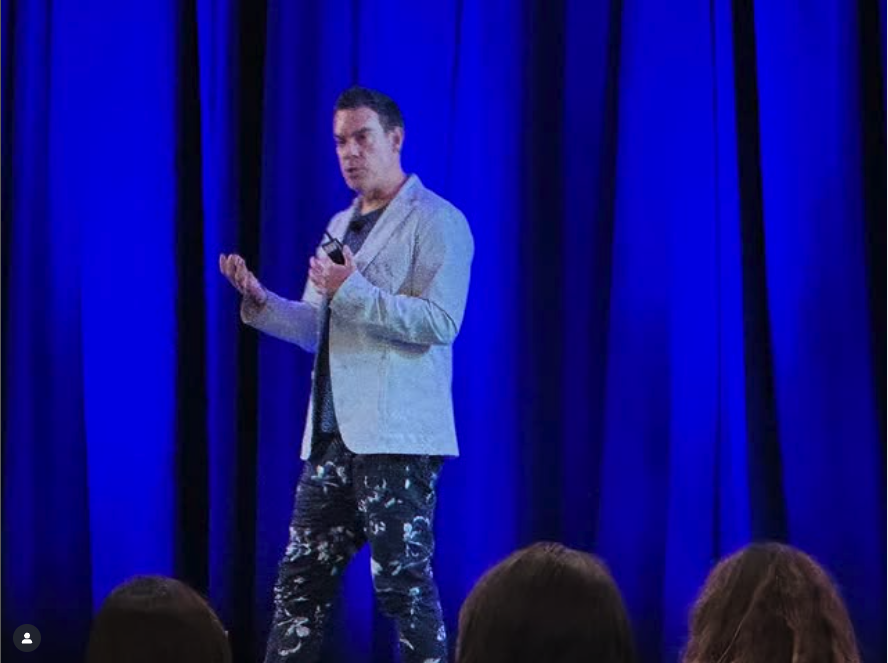 Man in a light-colored blazer and patterned pants speaking on stage with blue curtains in the background.