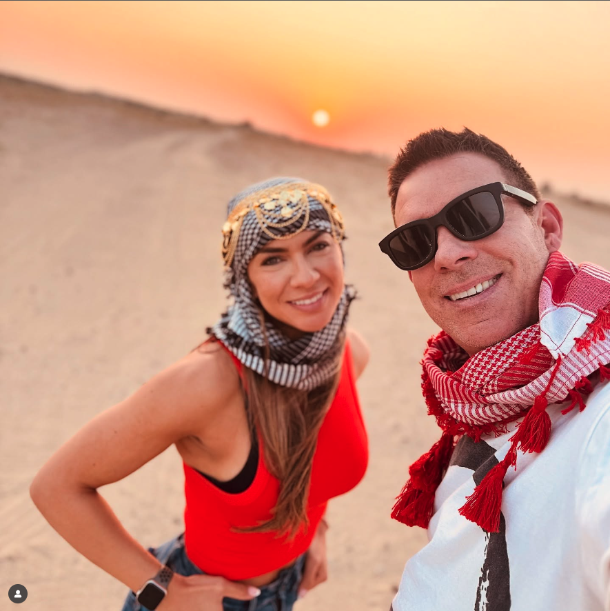 Smiling man and woman in traditional headscarves posing for a selfie in a sandy desert at sunset.