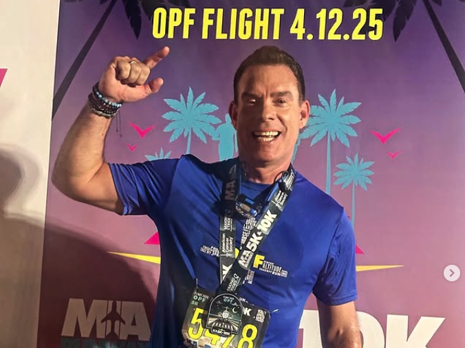 Smiling man wearing a blue shirt and race medals pointing upward at a backdrop with palm trees and text 'OPF FLIGHT 4.12.25'.