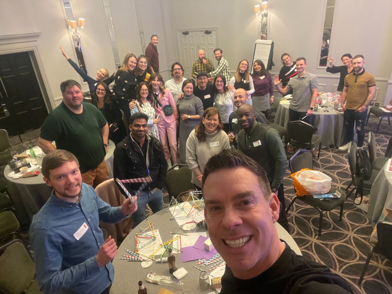 Group of diverse people smiling and posing for a selfie in a conference room with tables and chairs.
