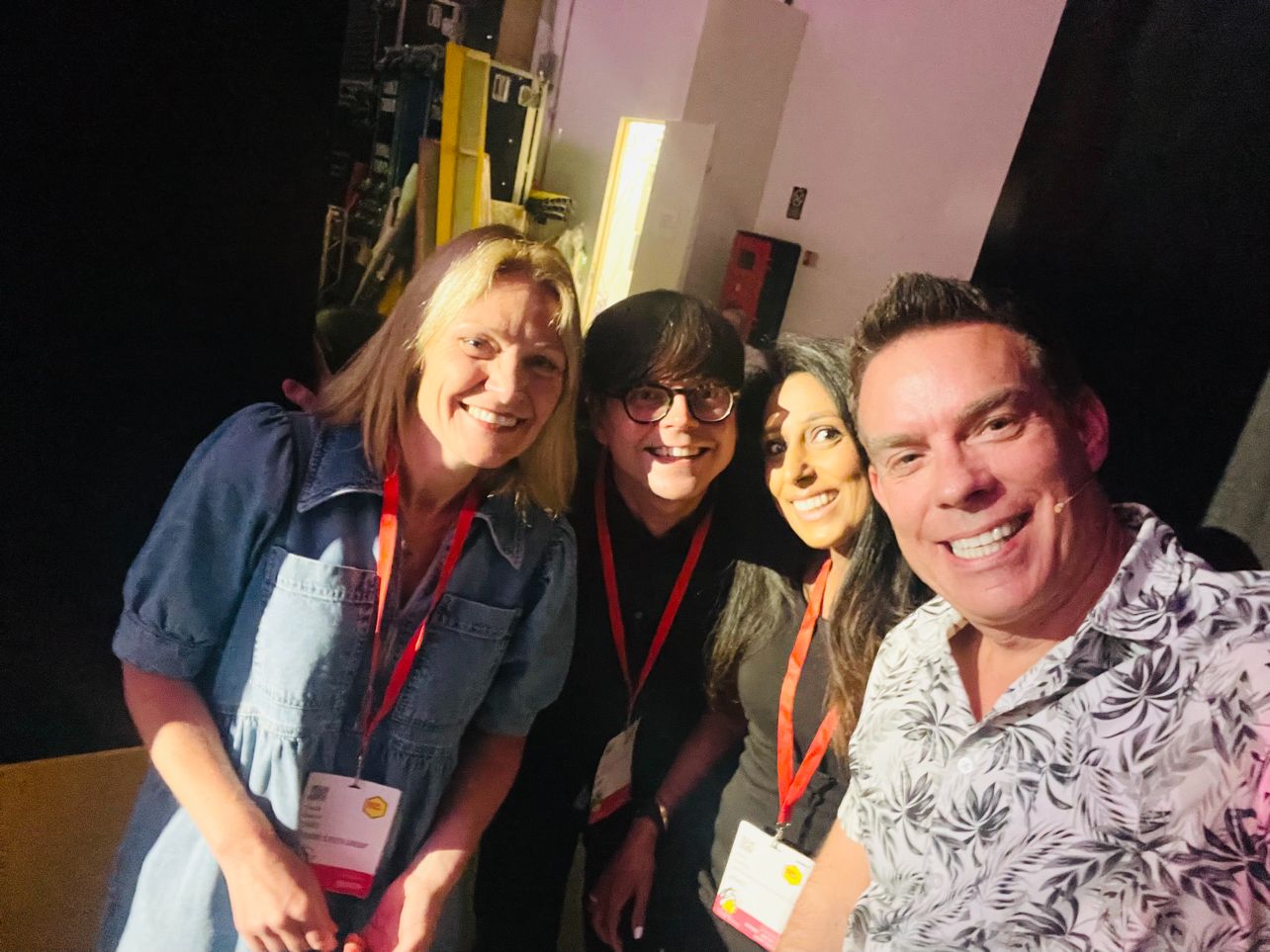 Four smiling people wearing event badges and red lanyards, posing together indoors with a dark background.