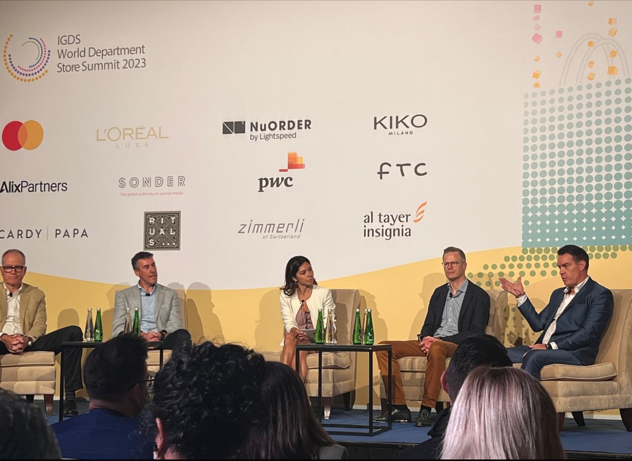 Panel discussion at IGDS World Department Store Summit 2023 with four men and one woman seated on stage with brand logos behind them.