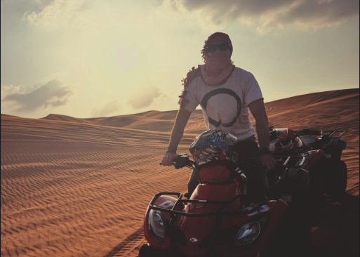 Person wearing sunglasses and a headscarf sitting on a red ATV in a desert with sand dunes at sunset.