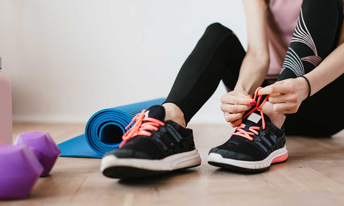 Person tying bright coral shoelaces on black running shoes sitting near a blue yoga mat and purple dumbbells on a wooden floor.