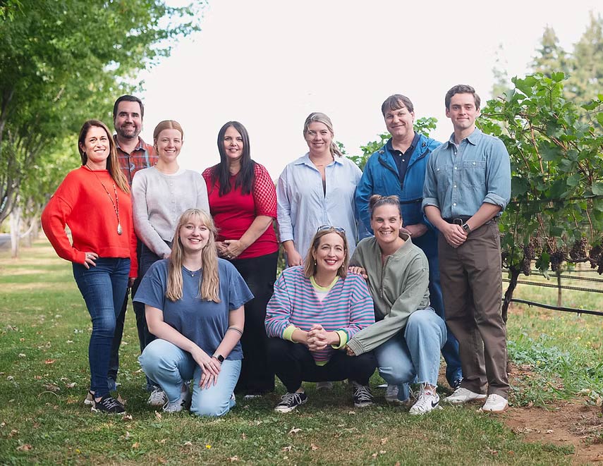 Group of ten adults smiling and posing outdoors on grass near grapevines and trees.