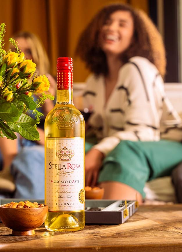 Bottle of Stella Rosa Moscato d'Asti wine on a wooden table with snacks and a blurred background of two women sitting and laughing.