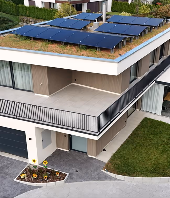 Modern house with solar panels on a green roof, a balcony with black railing, and a small garden with sunflowers.