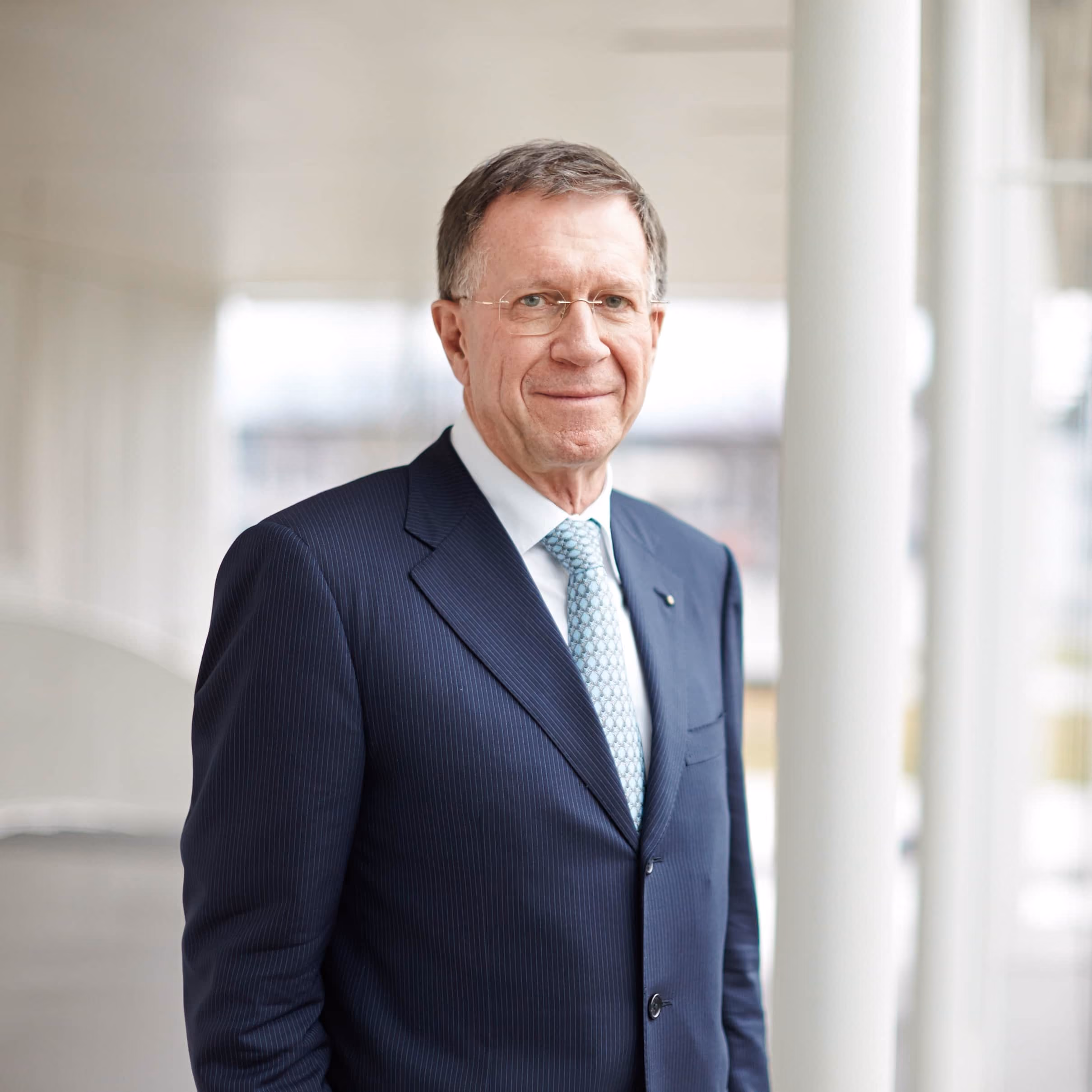 Älterer Mann mit Brille im dunkelblauen Nadelstreifanzug und hellblauer Krawatte vor hellem, modernem Hintergrund.