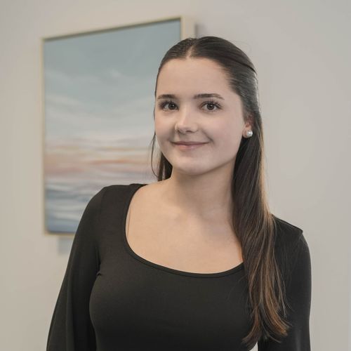 Junge Frau mit langen braunen Haaren, schwarzem Oberteil und Perlenohrringen lächelt vor einem Bild mit sanften Farben.