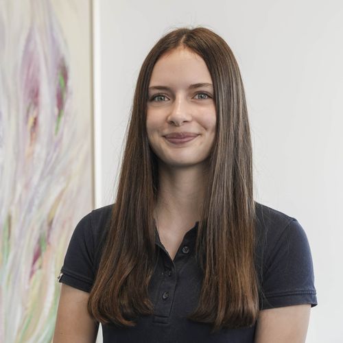 Junge Frau mit langem braunem Haar und dunkelblauem Poloshirt, die leicht lächelt vor einem hellen Hintergrund mit abstraktem Gemälde.