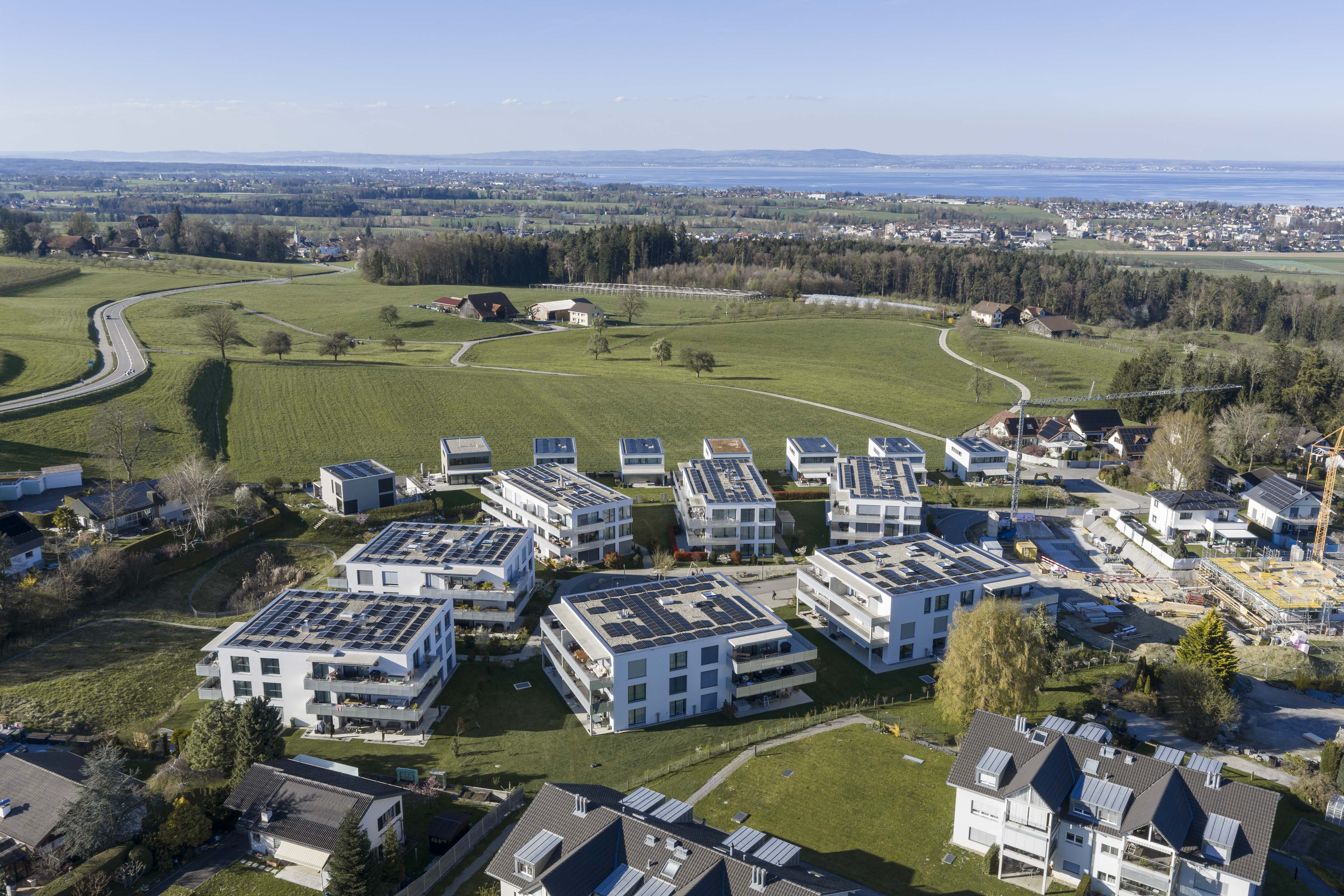 Luftaufnahme einer Wohnsiedlung mit modernen, weißen Mehrfamilienhäusern mit Solarpaneelen auf den Dächern, umgeben von grünen Feldern und weiter Landschaft.