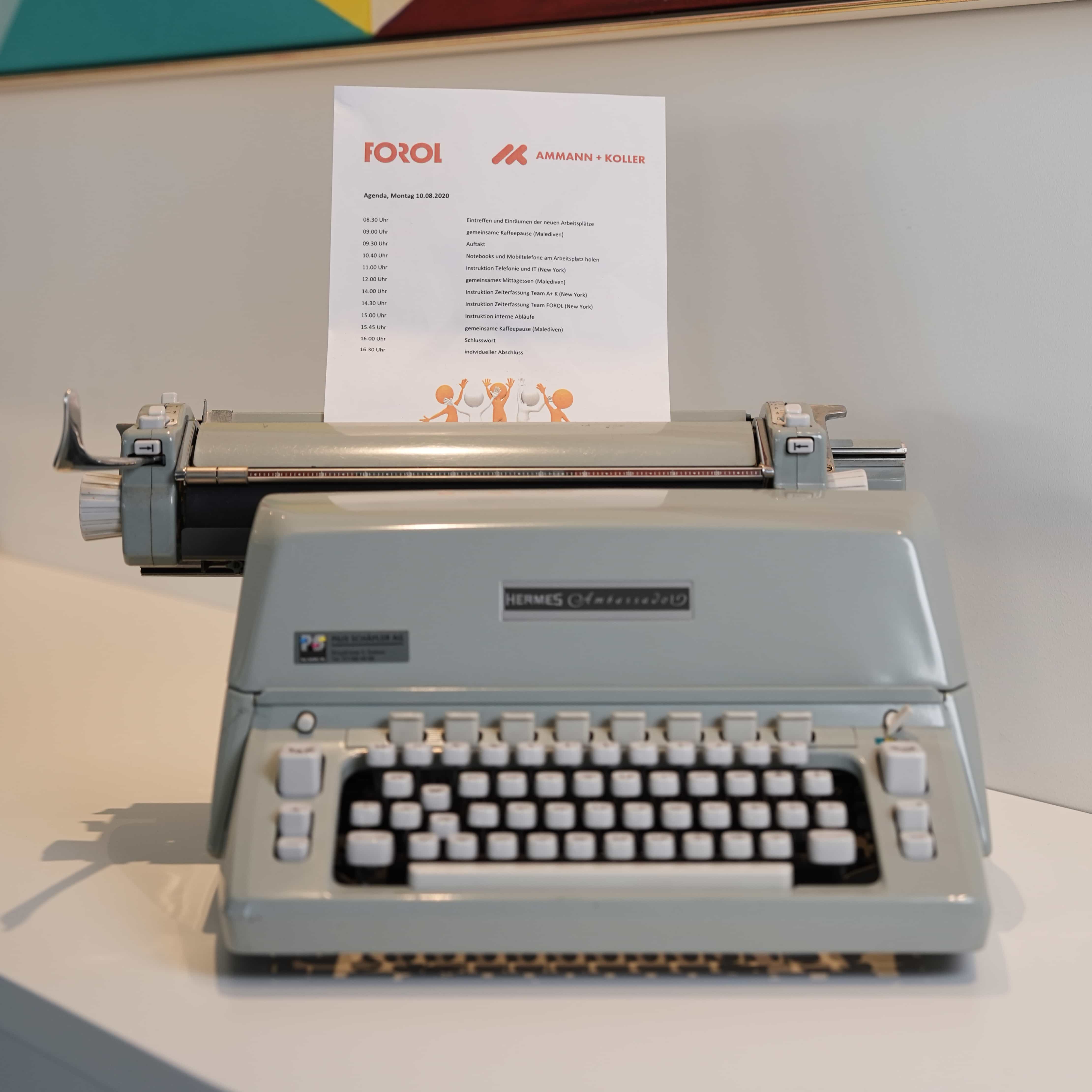 Eine graue Hermes Ambassador Schreibmaschine mit einem Blatt Papier, auf dem eine Agenda in deutscher Sprache steht.
