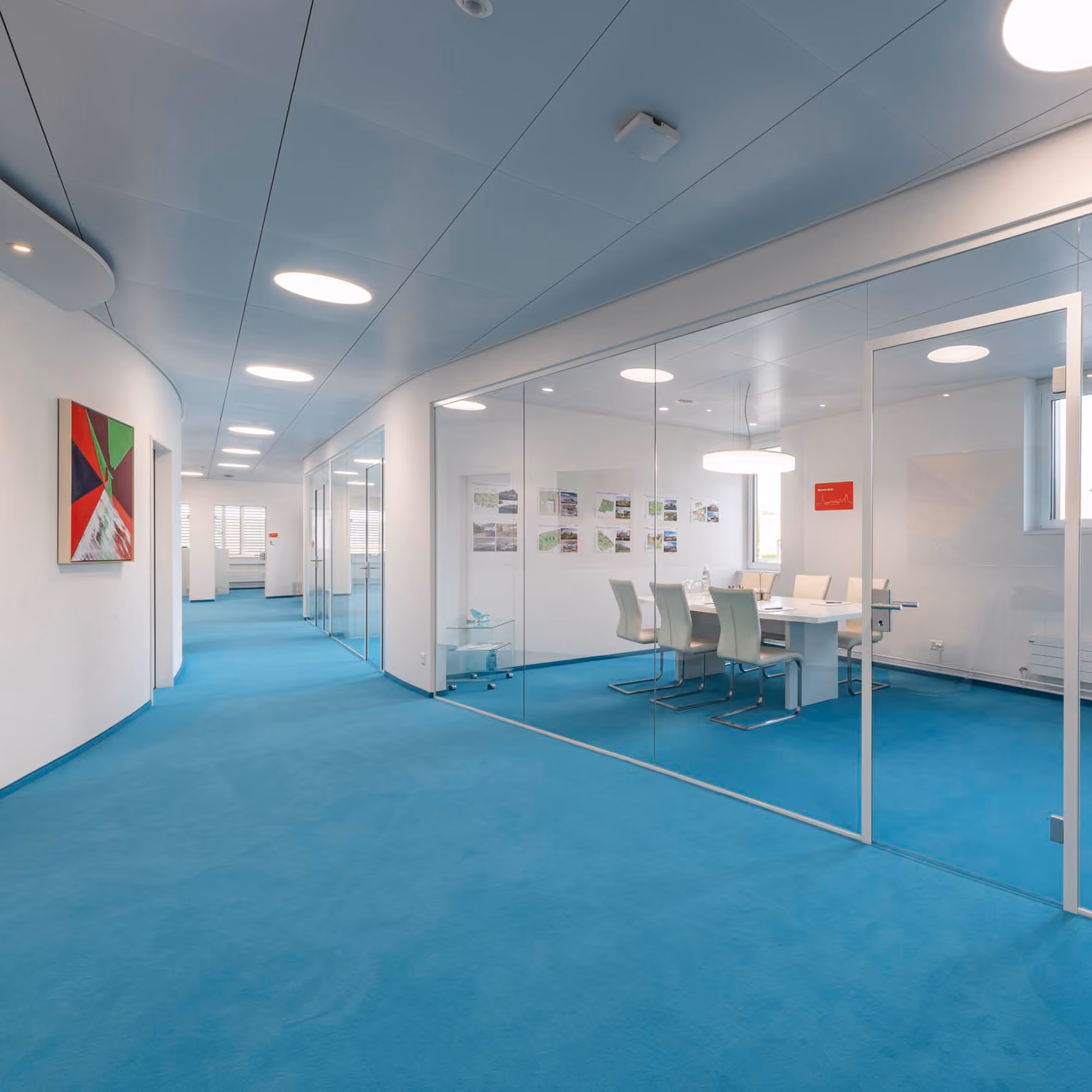 Moderner Büroflur mit blauem Teppich, weißen Wänden, Glaswänden und einem Besprechungsraum mit weißen Stühlen.