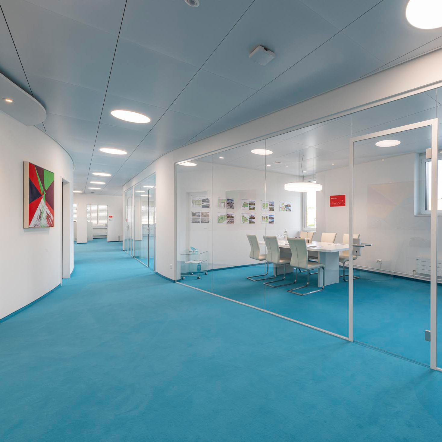 Moderner Büroflur mit blauem Teppich, weißen Wänden, Glaswänden und einem Besprechungsraum mit weißen Stühlen.