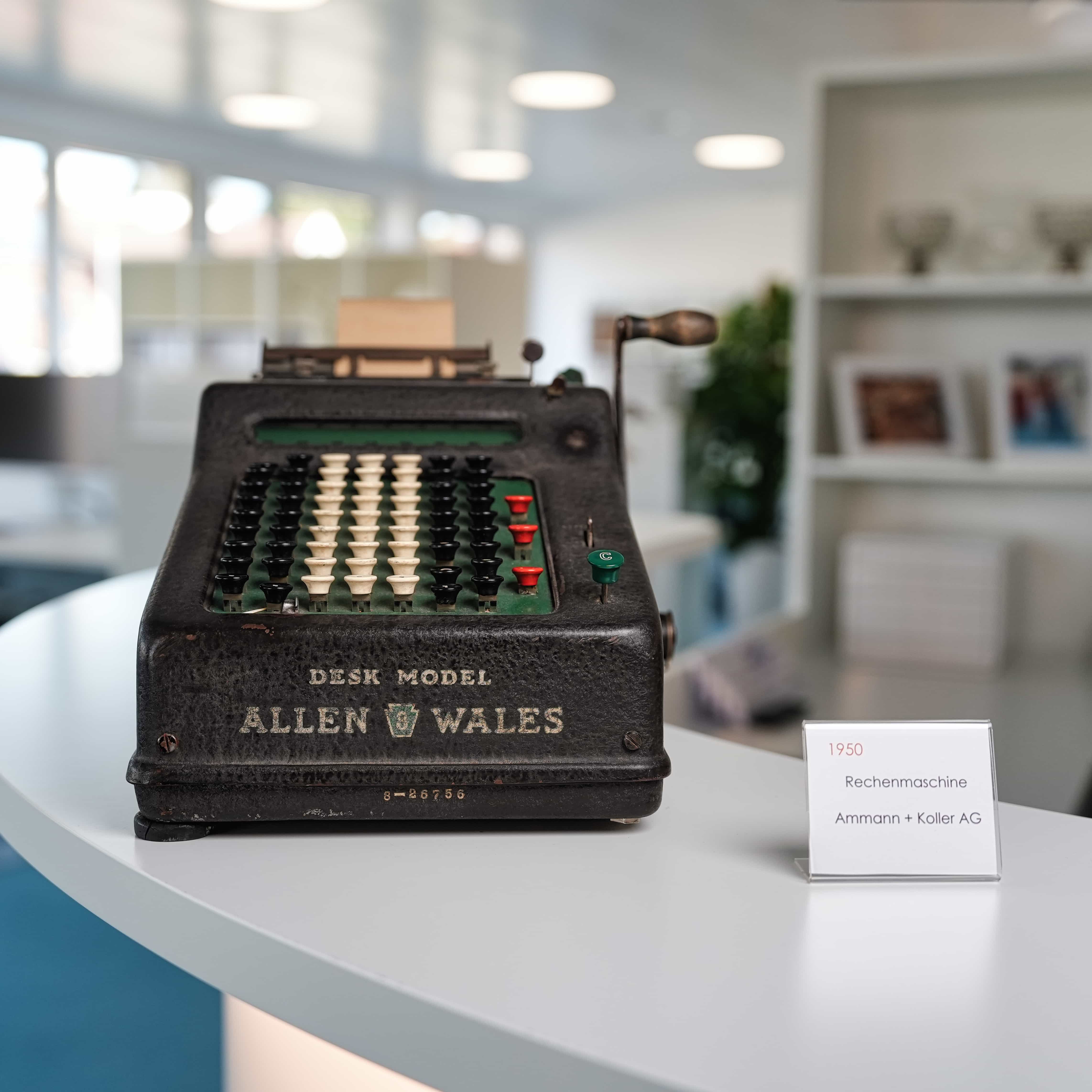 Vintage mechanische Rechenmaschine Modell Allen Wales aus dem Jahr 1950 auf einem weißen Tisch mit einer kleinen Informationstafel.
