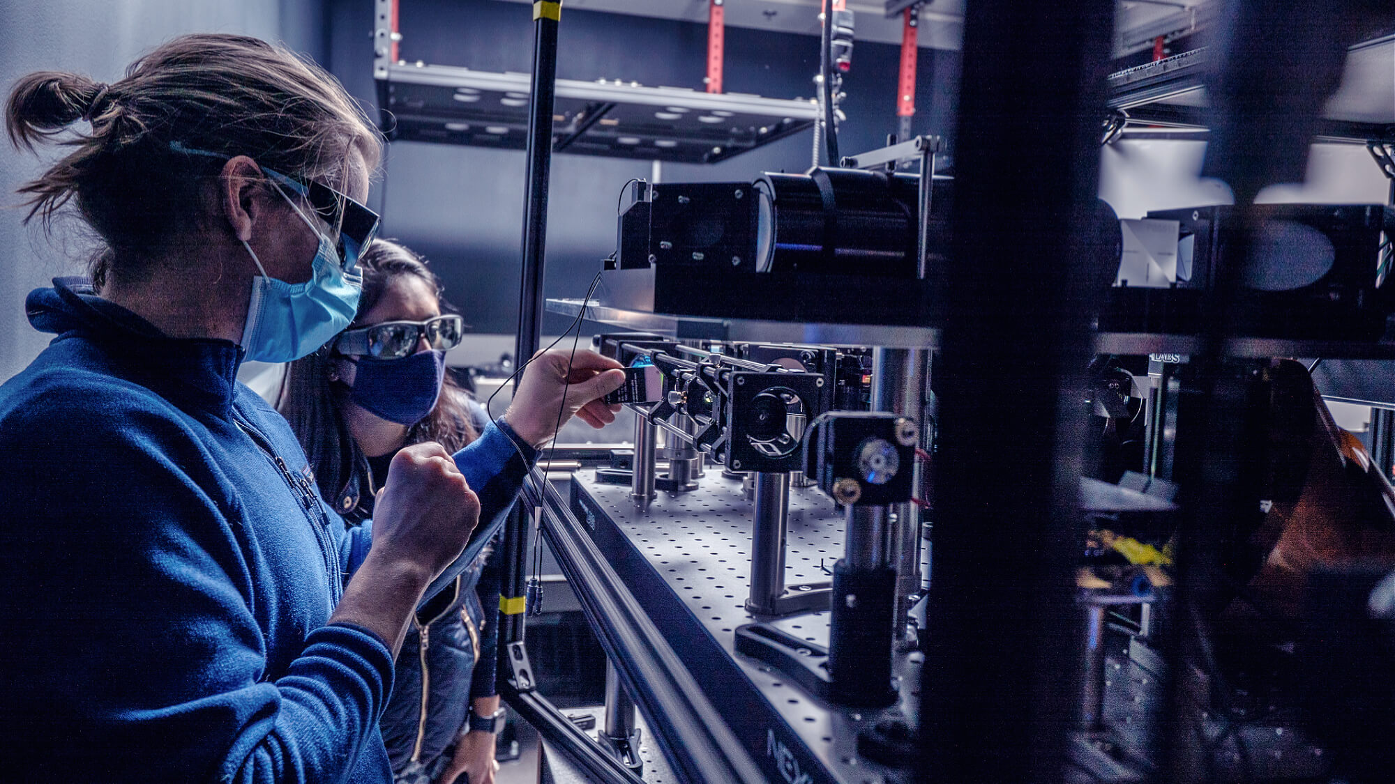 Scientists in lab coats and masks operating advanced optical laboratory equipment with multiple machines