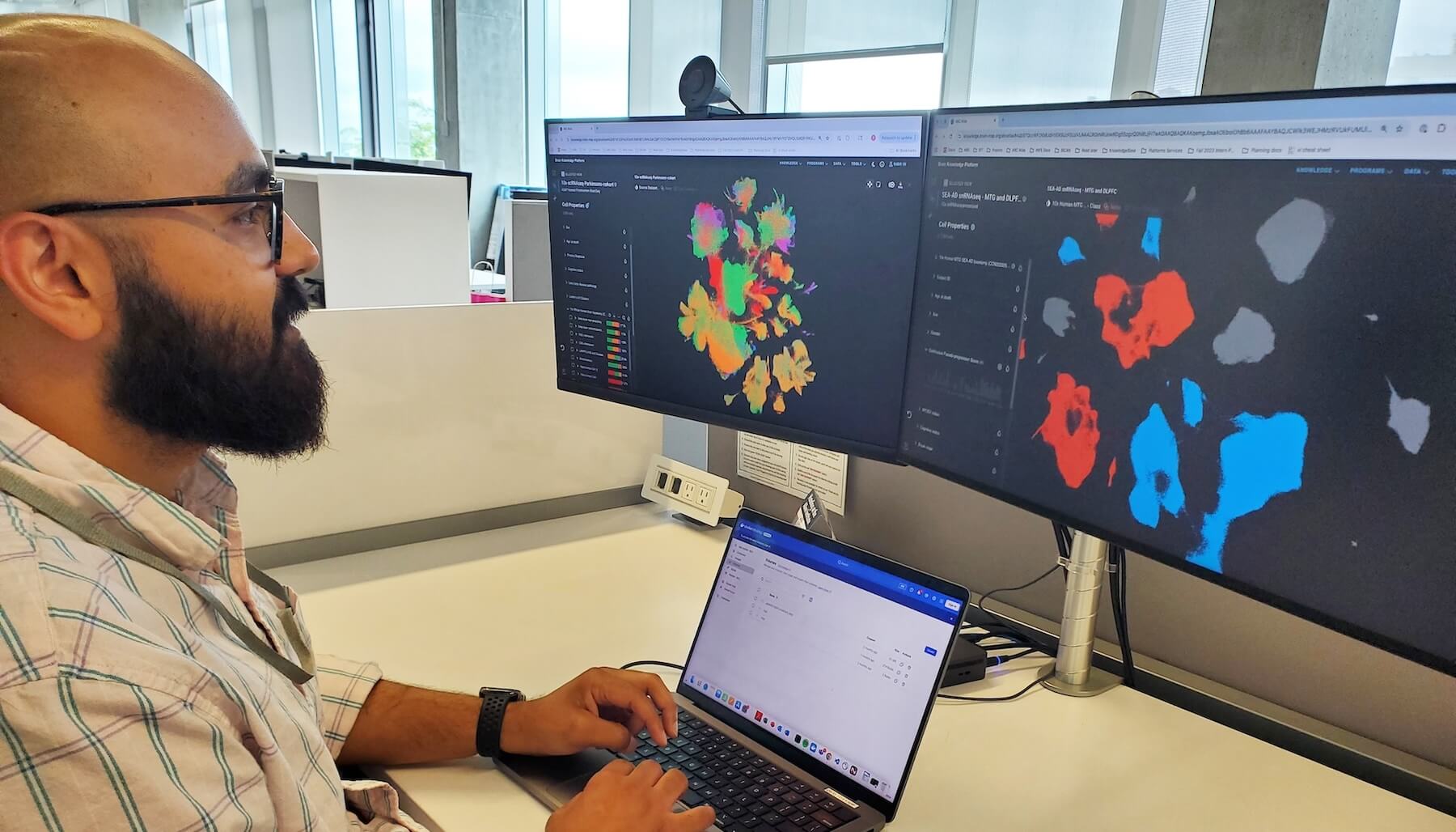 Man with glasses works at laptop viewing colorful data visualizations on dual monitors in office.