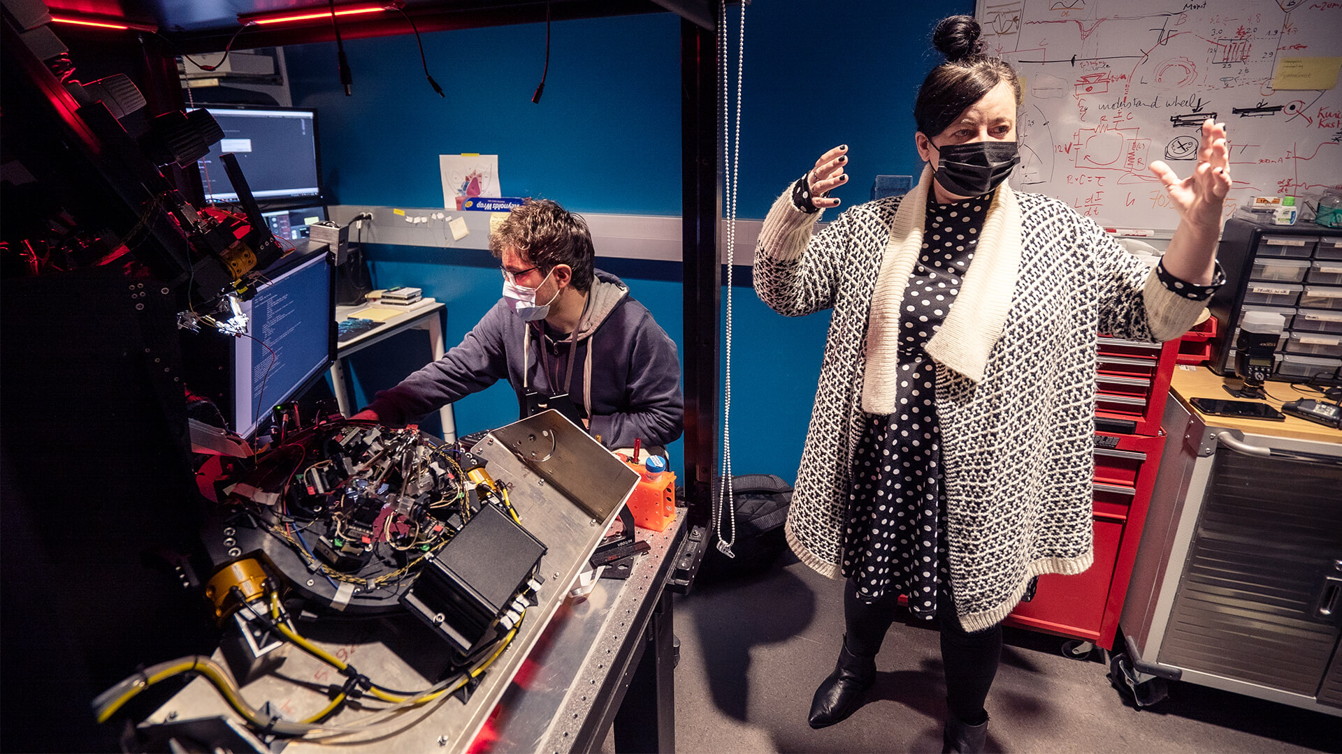 Two masked people in a high-tech lab with electronics, red neon lighting, and whiteboards.