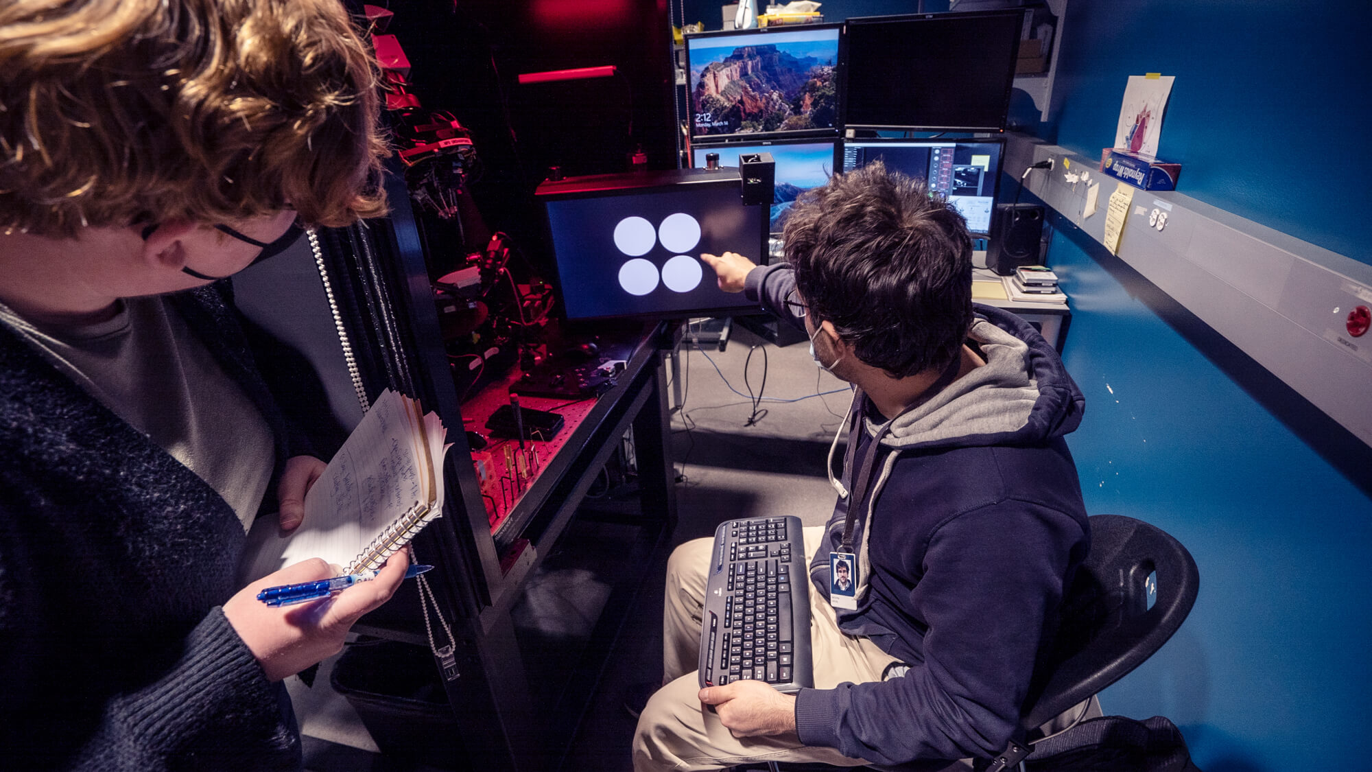 Two people work at a high-tech control station with multiple monitors and red lighting.