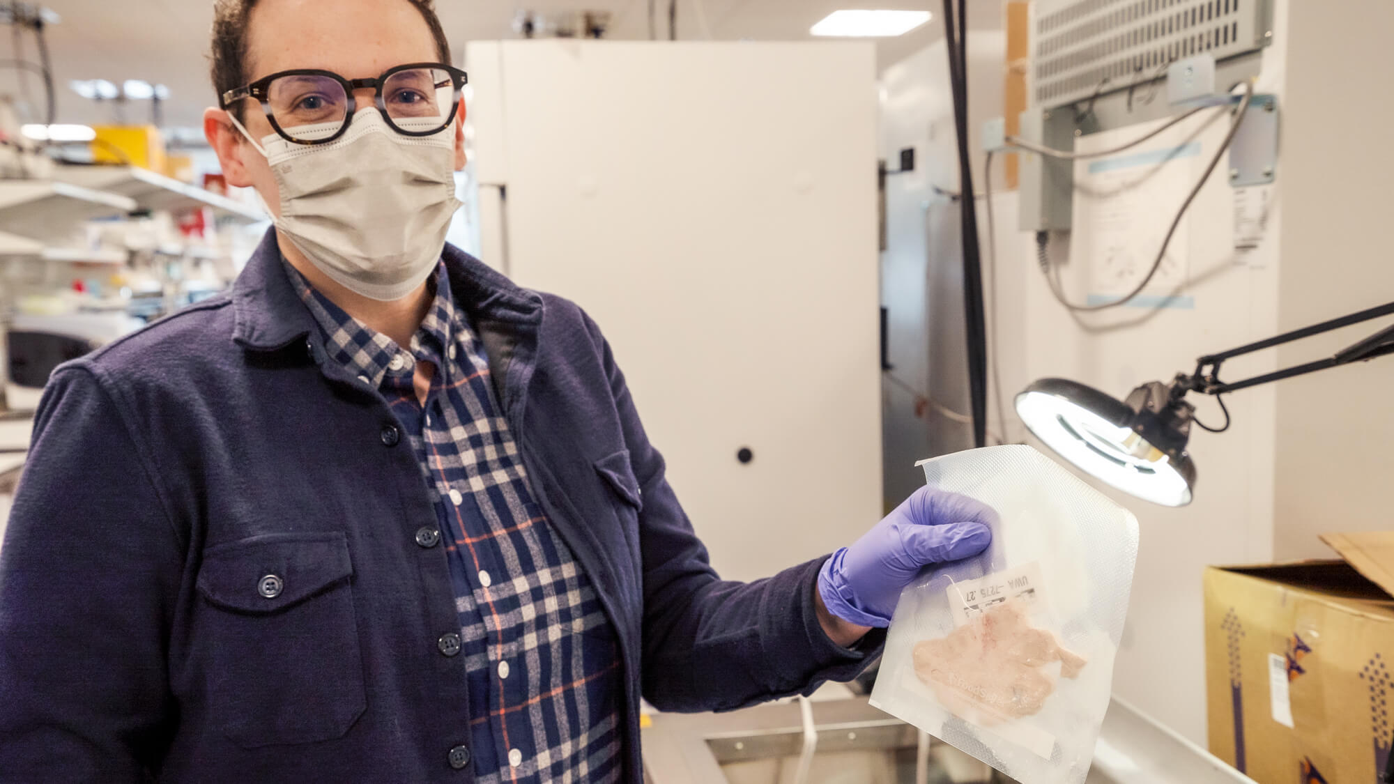Scientist in lab coat, mask, and glasses examining sample in laboratory.