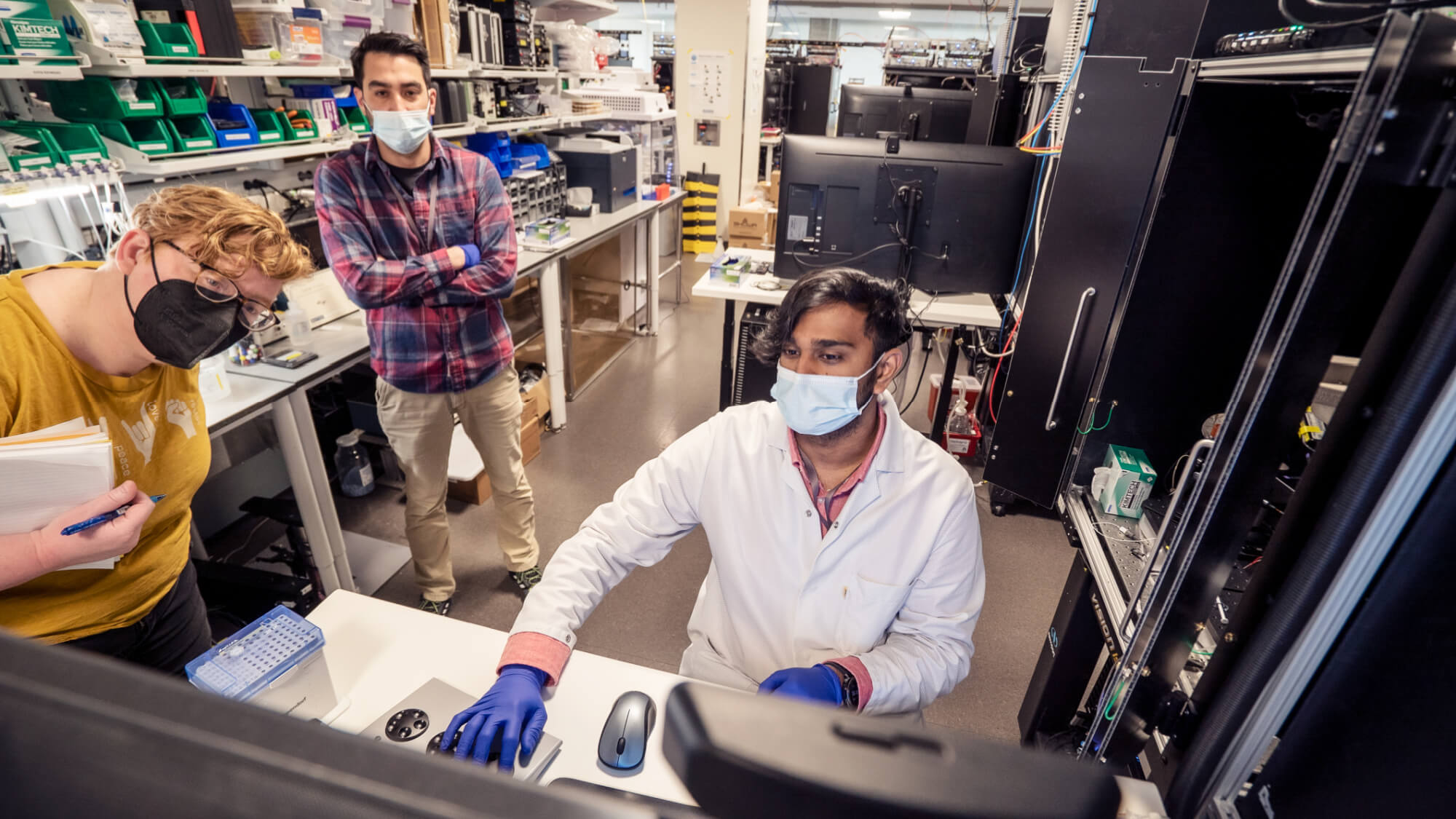 Three masked scientists work in a laboratory with computer equipment and shelves of supplies