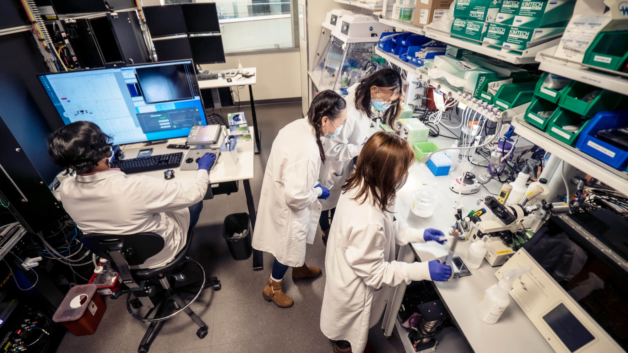 Three scientists in white lab coats working in a modern laboratory with equipment and computers.