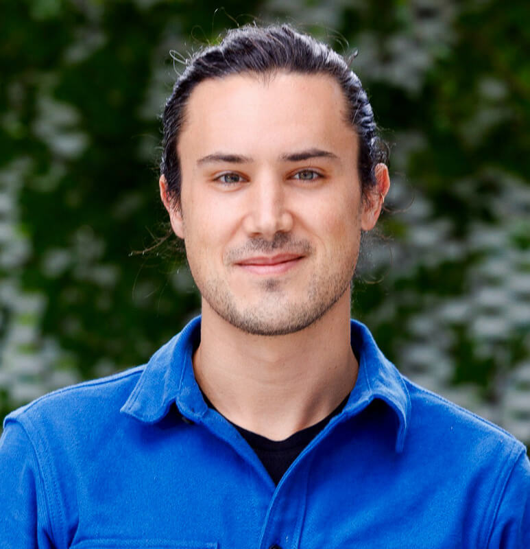 Man with dark hair wearing blue jacket, green foliage background, professional headshot portrait.