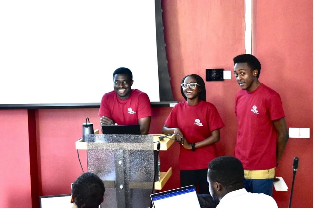 Three smiling young people in red t-shirts present at podium against pink wall background.