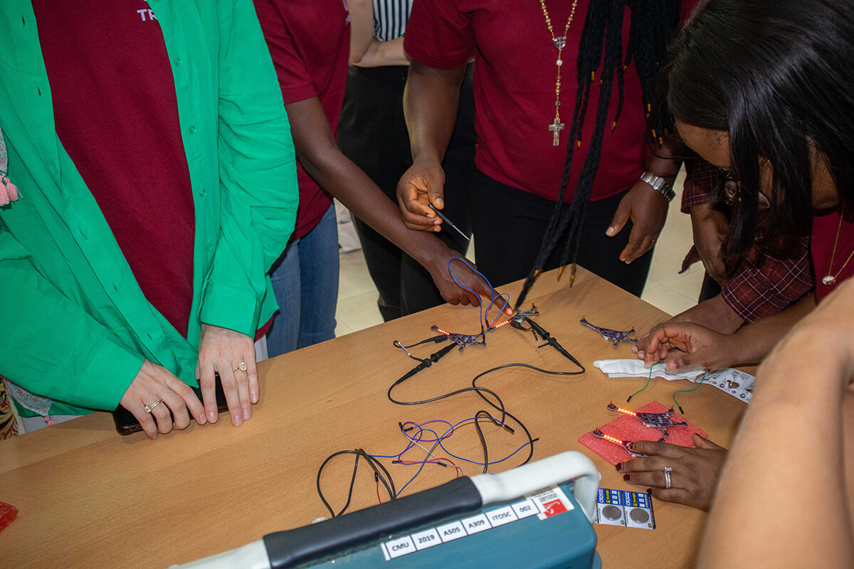 Group of people in burgundy shirts assembled circuit boards and wires on wooden table together.