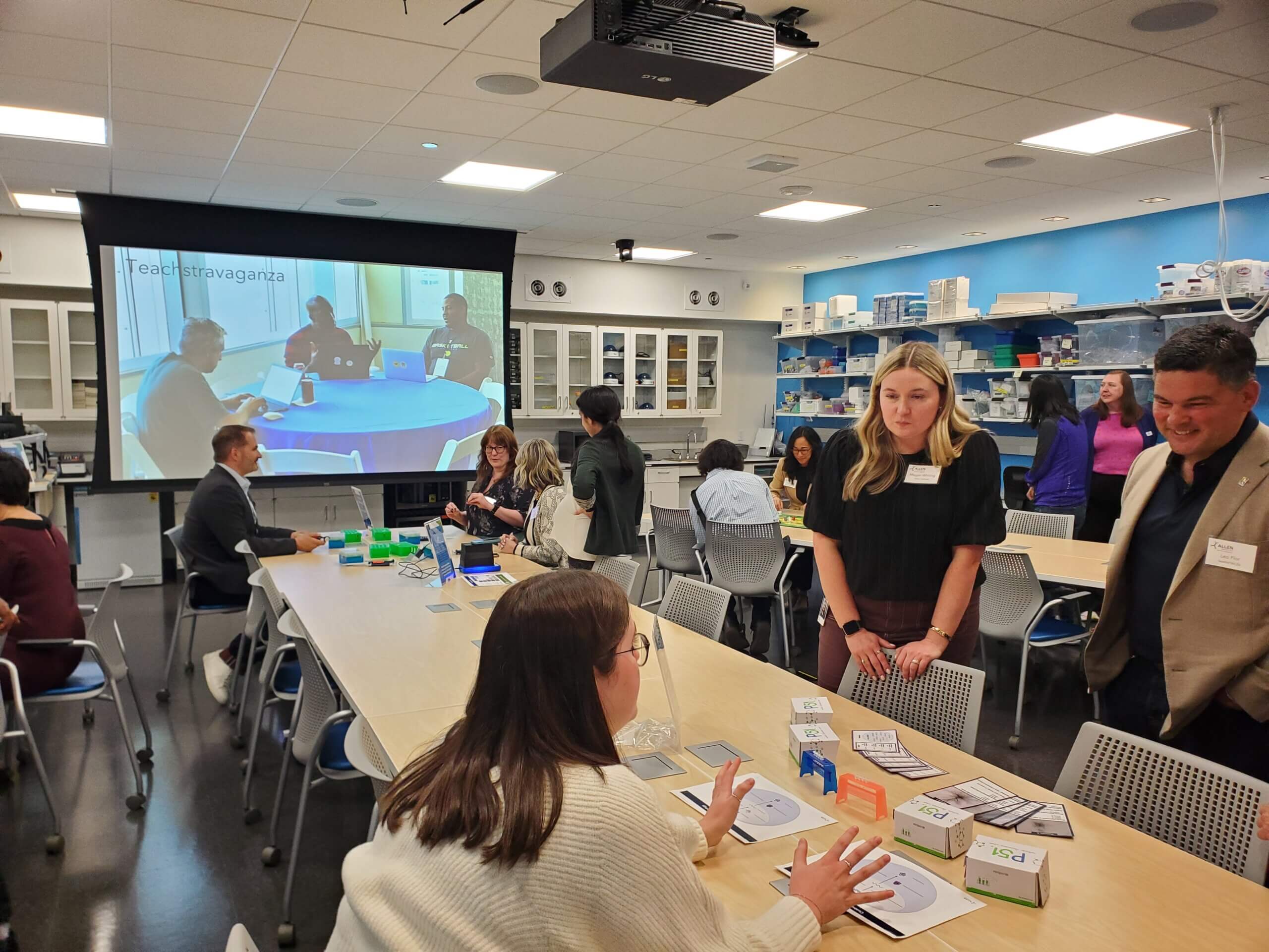 Educational workshop in classroom with attendees watching presentation on projected screen and tables with teaching materials.