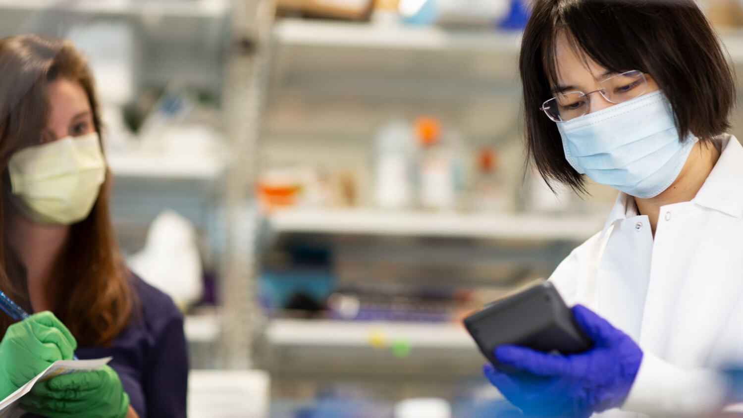 Scientist in mask and gloves holding tablet in laboratory with colleague nearby