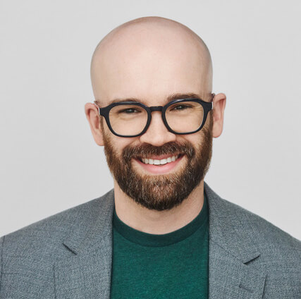 Smiling bald man with dark beard wearing glasses and gray blazer