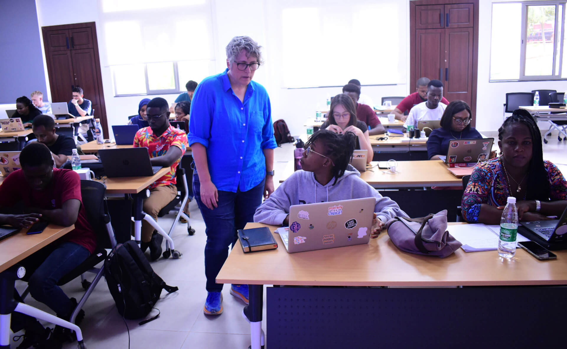 Instructor in blue shirt teaching diverse group of students using laptops in bright classroom.