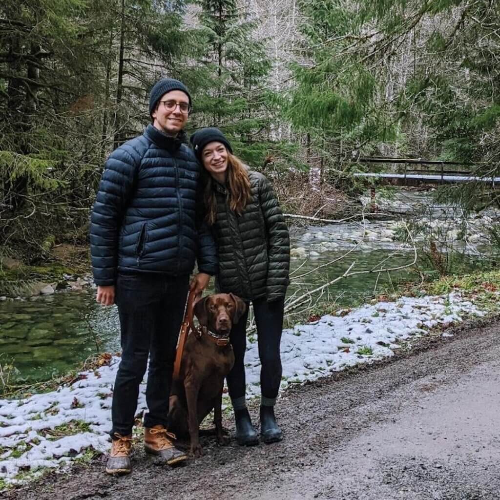 Couple with brown dog posing on forest path by snowy stream and bridge.