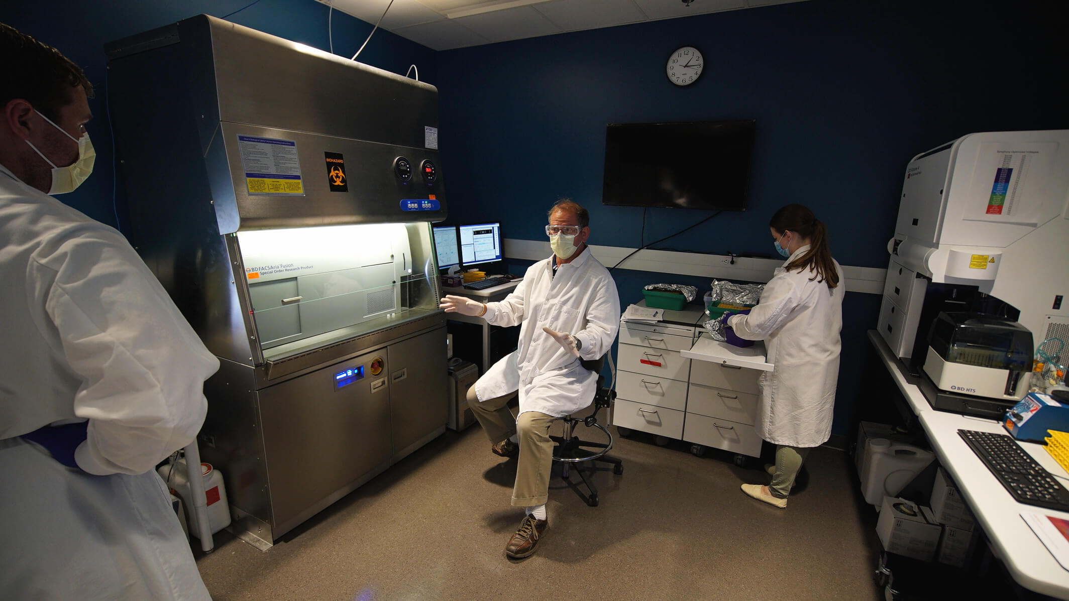 Laboratory technicians in white coats and masks working in biosafety cabinet room with medical equipment