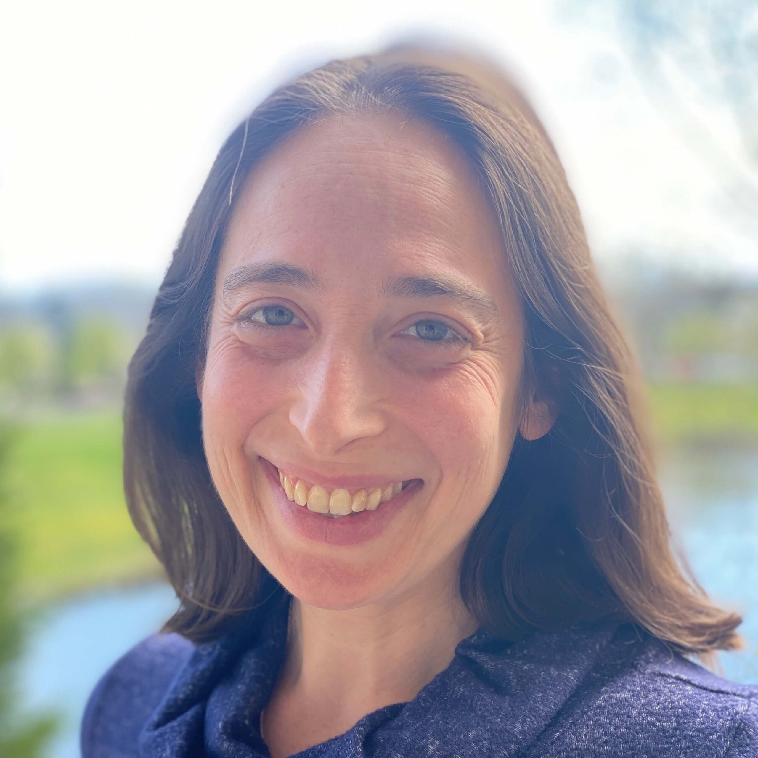 Portrait of a smiling woman with brown hair outdoors, blurred green landscape background.