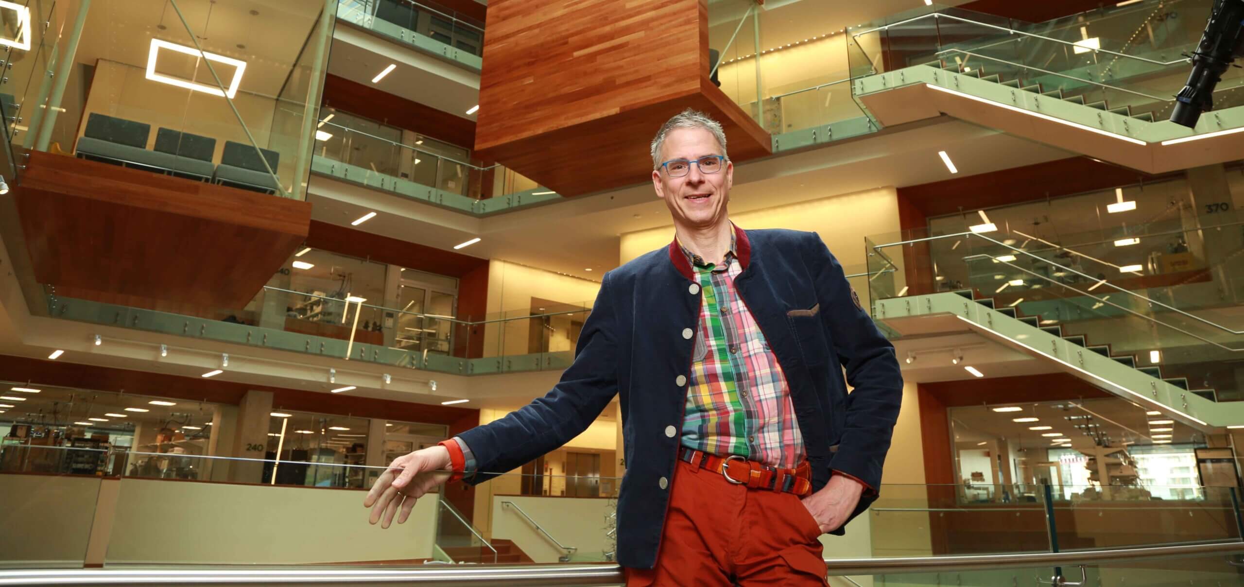 Man in navy blazer and red pants stands in modern multi-story atrium with glass railings.