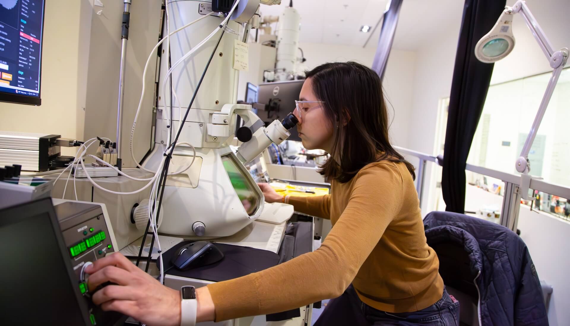 Scientist in yellow sweater using microscope in laboratory with equipment and monitors.