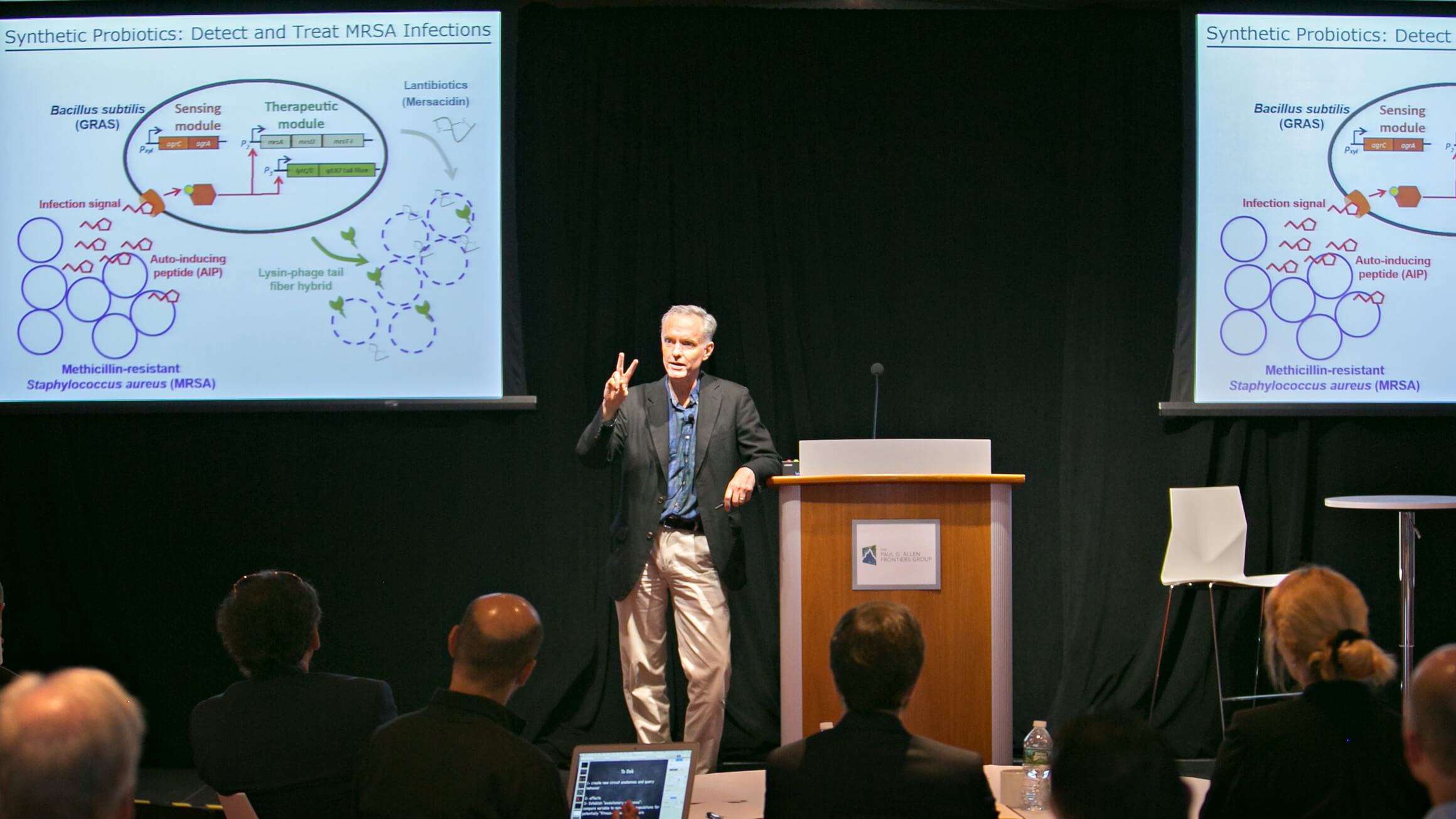 Scientist presenting research on synthetic probiotics for MRSA infection detection and treatment at podium.