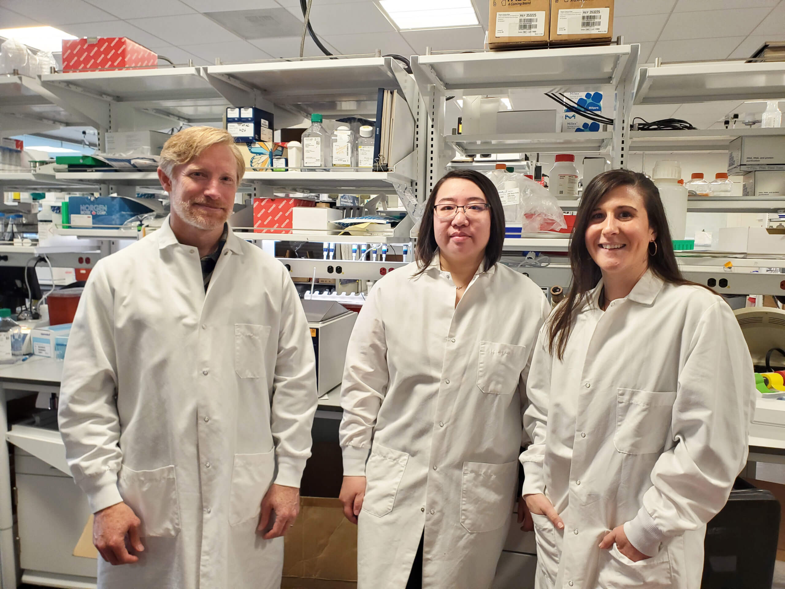 Three scientists in white lab coats standing together in a laboratory with shelves of equipment and supplies.