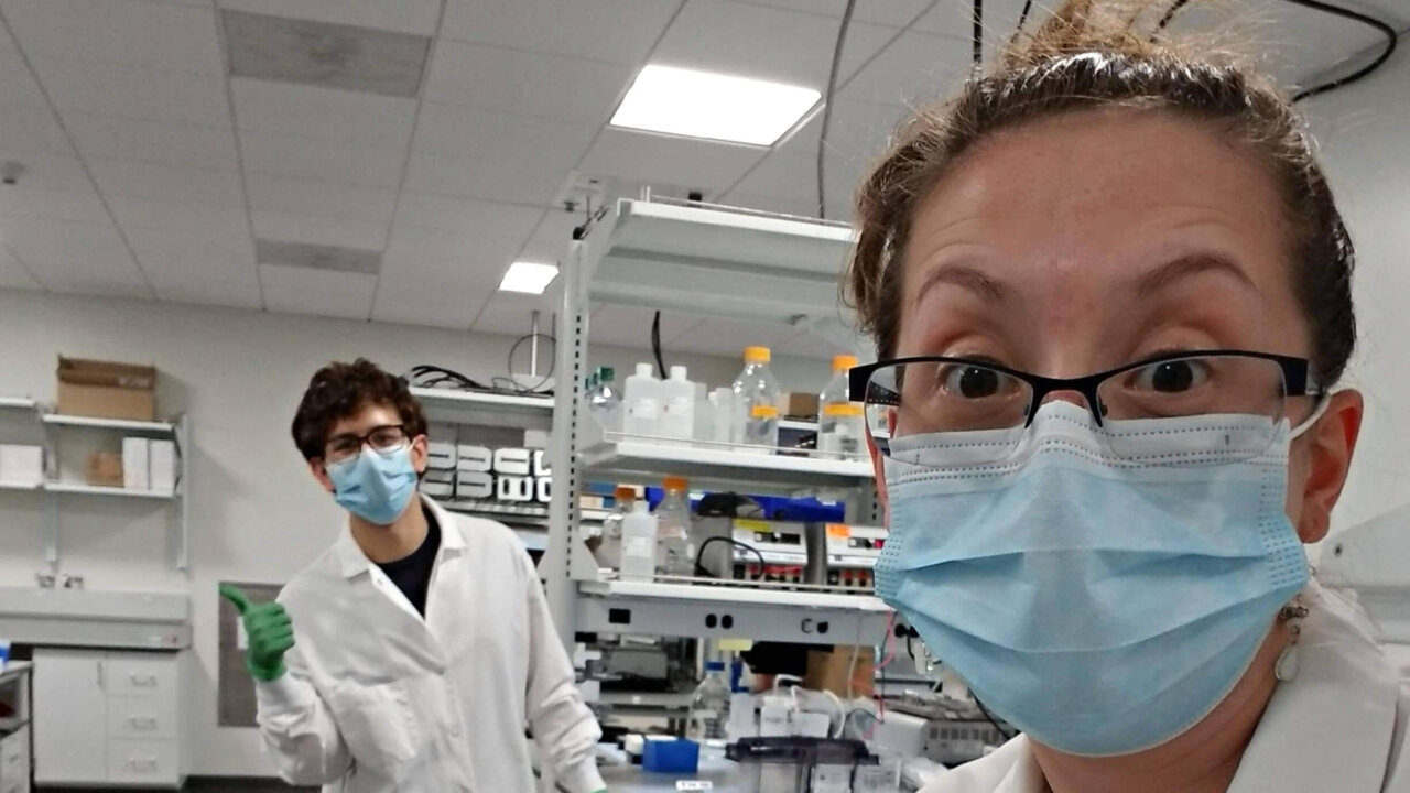 Two lab technicians wearing masks and white coats in a laboratory with equipment and supplies.