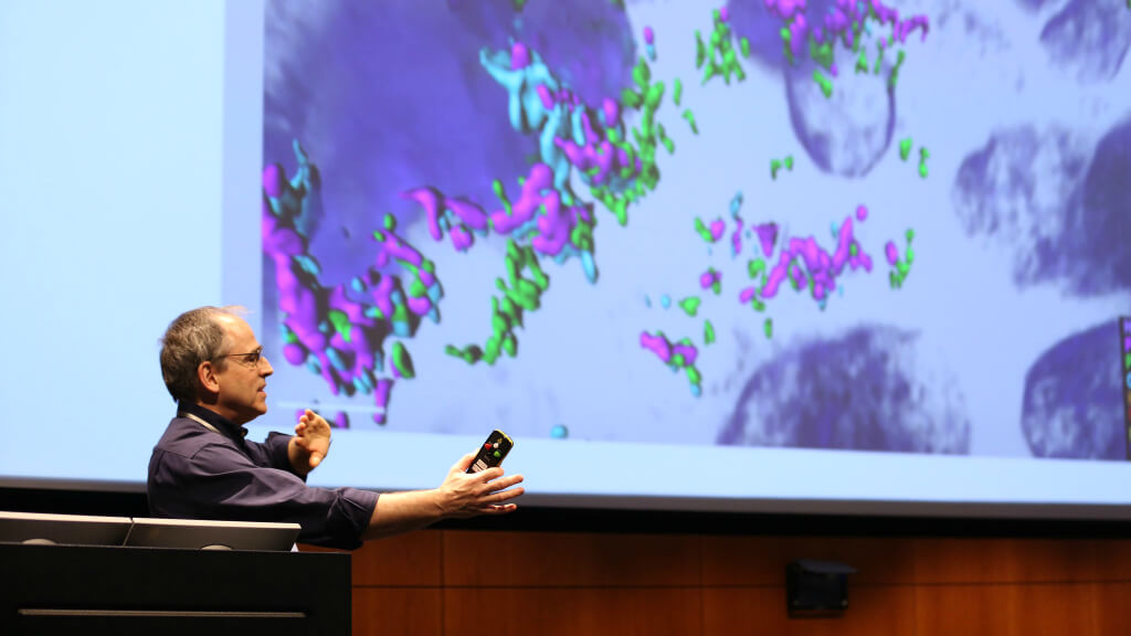 Man presenting at podium with colorful molecular visualization on screen behind him