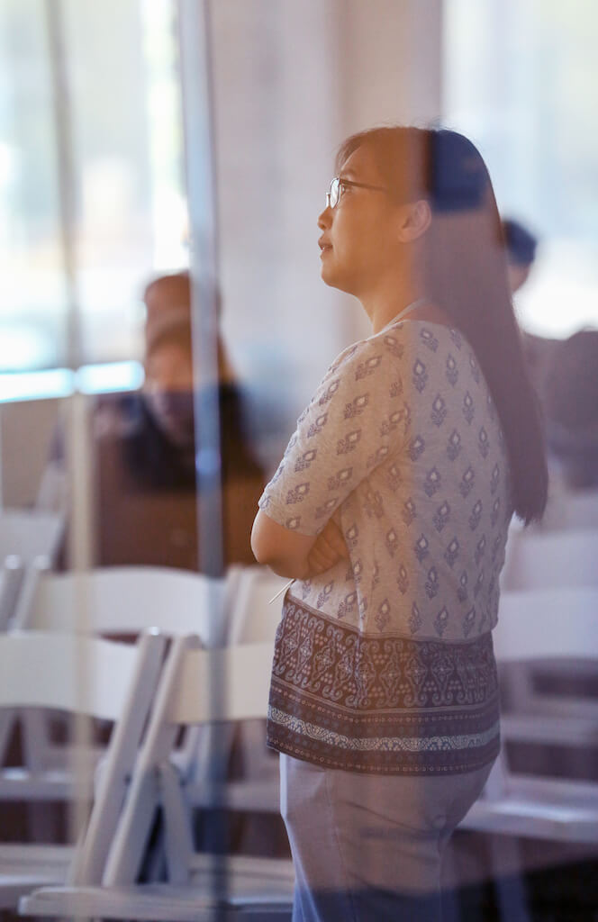 Woman wearing glasses standing by window in bright, blurred indoor setting.