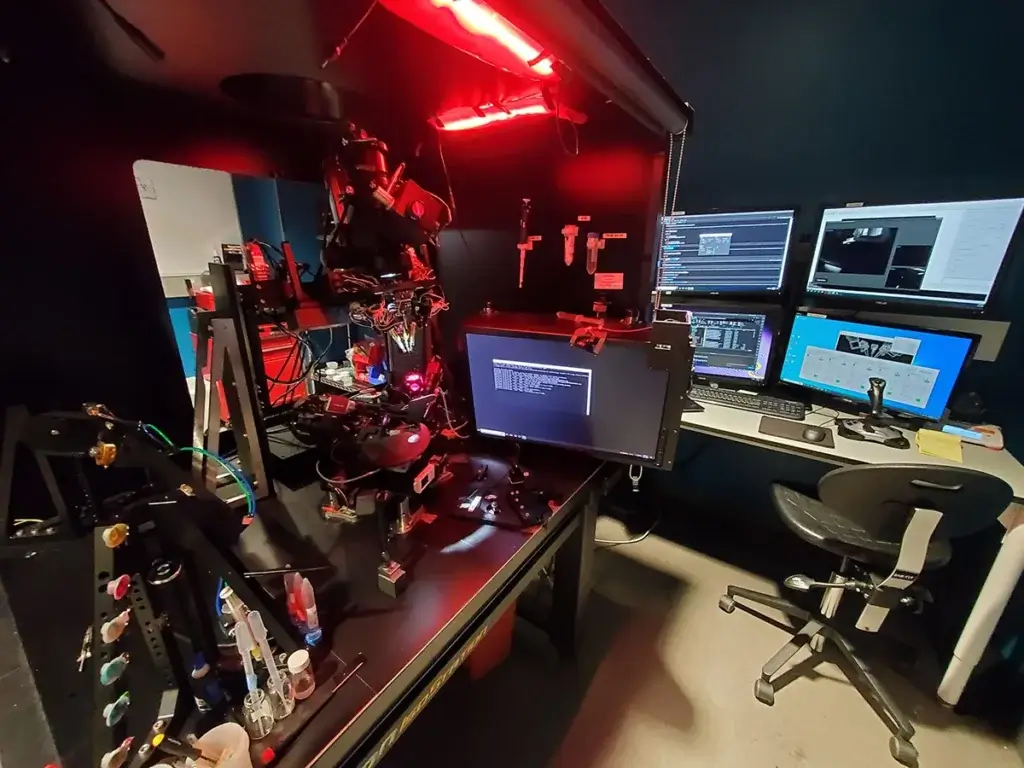 High-tech workspace with 3D printer under red lighting and multiple computer monitors displaying software interfaces.