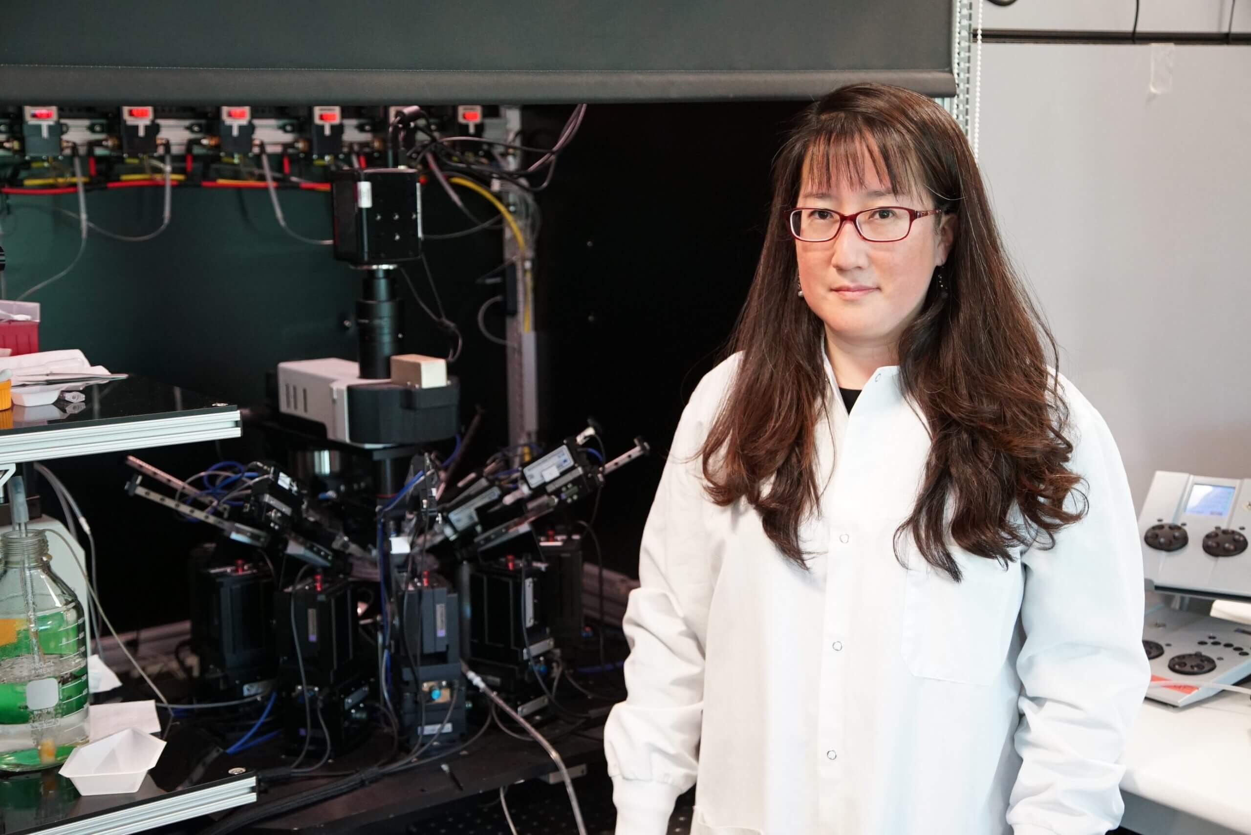 Photo of a woman in a white lab coat standing next to a large electrophysiology rig