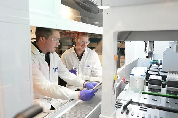 Two scientists in lab coats work with equipment inside a biosafety cabinet in laboratory.
