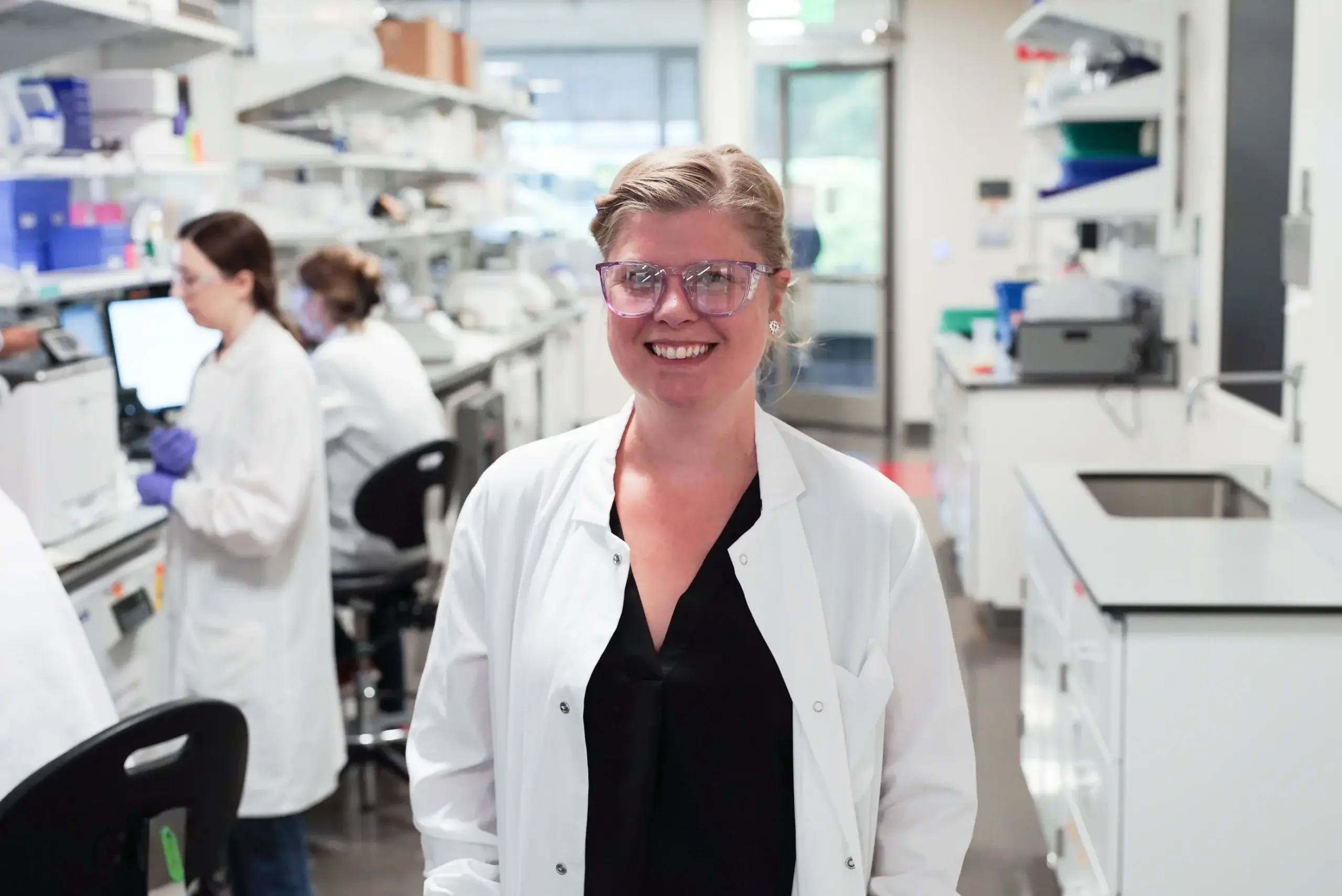 Smiling female scientist in white lab coat and glasses in modern laboratory with colleagues and equipment